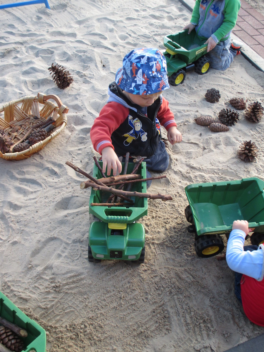 Children playing with trucks, sticks and pine cones in the babies sandpit